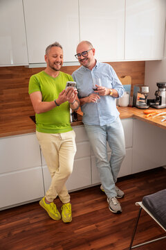 Happy two gay men standing together in the kitchen while sharing something on a mobile phone