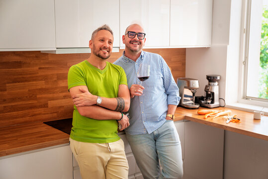 Happy male couple standing in the kitchen with a glass of wine, smiling directly at the camera