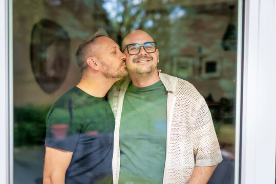 Two gay men standing behind a window in the living room, one kissing the other