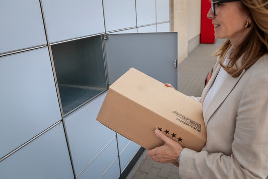 A middle-aged woman holding a parcel while standing in front of a parcel locker