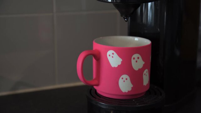 Hand places a bright pink Halloween ghost mug on a coffee machine, showing seasonal lifestyle coffee use in a home, office or hospitality setting.