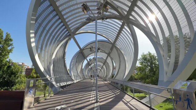 First person perspective of a person walking across the modern Arganzuela footbridge, a famous spiral-shaped landmark in Madrid R&iacute;o Park, during a sunny day in the capital city of Spain