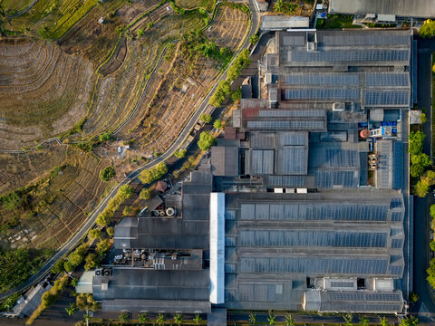 Aerial view of a large industrial factory complex with extensive solar panel arrays on the roof, situated next to terraced rice fields and a road in Semarang Regency, Central Java, Indonesia.