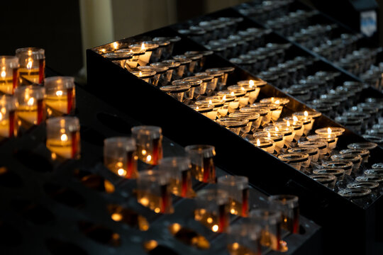 Rows of small votive candles glowing in transparent holders on tiered stands, creating a warm and atmospheric scene in a dark interior.