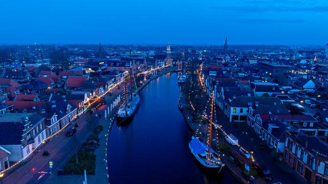 Aerial view of the canal in Lemmer at twilight, featuring historic buildings, traditional sailing ships, and festive string lights along the waterfront. Lemmer, Friesland, Netherlands.