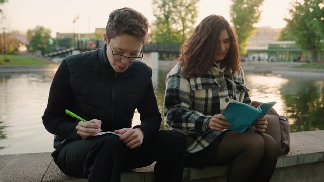 Two people sit by pond. One person writes with green pen while other reads from blue book. scene is set outdoors near water and trees notebook writing