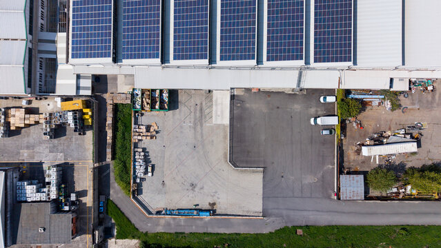 Aerial view of a manufacturing plant featuring large solar panel arrays on the roof, a paved loading area with parked delivery vehicles, and industrial storage zones in Milan, Lombardy, Italy.
