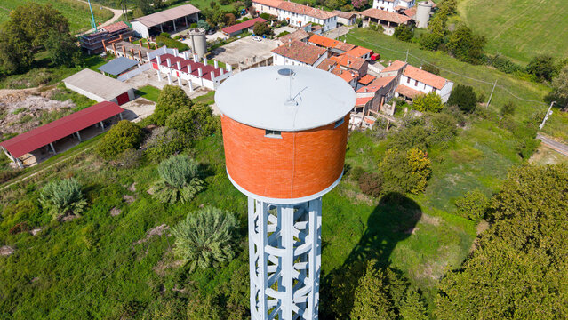 Aerial view of a water tower with a brick-colored tank and concrete support structure, surrounded by farm buildings, lush green fields, and trees in Mediglia, Lombardy, Italy.