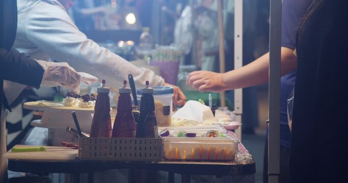 A gloved street food vendor prepares desserts beside condiment bottles while a customer reaches for their order at a Thai night market stall.