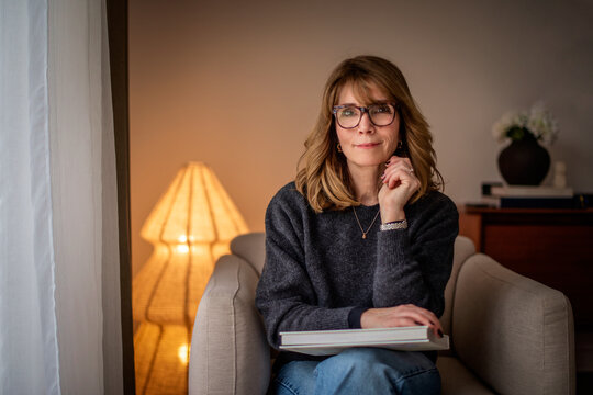 Confident middle-aged woman sitting indoors at home, reading a book