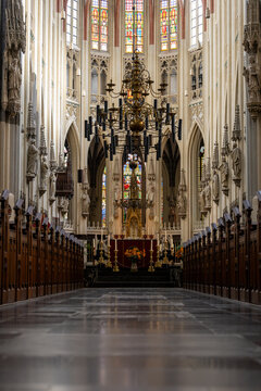 Vertical symmetrical view through an ornate Gothic church toward the altar, with tall stone columns, wooden choir stalls, chandeliers, religious statues and colorful stained glass windows.