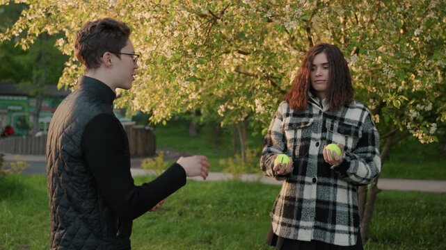 Man wearing glasses stands facing another man under blossoming trees. second subject dressed in checkered coat juggles tennis balls while first man watches and gestures