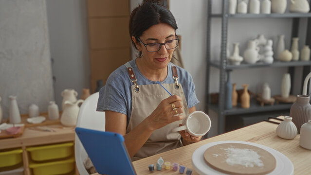 Woman painting ceramic mug, holds handle and paints with brush in studio wearing apron and using a tablet for reference; calm focus.