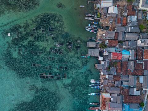 Aerial view of Karimunjawa village with traditional fishing boats moored along the coast and fish farming cages in the clear turquoise water, Karimunjawa, Central Java, Indonesia.