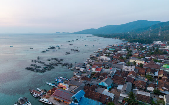 Aerial view of Karimunjawa village with traditional houses, fishing boats in shallow turquoise waters, and lush mountains in the background in Karimunjawa, Central Java, Indonesia.
