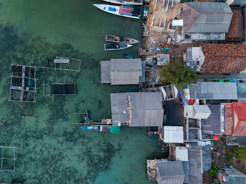 Aerial view of Karimunjawa featuring stilt houses over turquoise water, wooden fishing boats, and fish farming nets along the coast. Karimunjawa, Central Java, Indonesia.