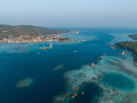 Aerial view of Karimunjawa islands featuring turquoise coral reefs, small boats, and a coastal village nestled against lush green hills in Karimunjawa, Central Java, Indonesia.