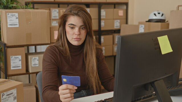 Woman holding creditcard with hand toward computer monitor amid parcel boxes in building; online order frustration.