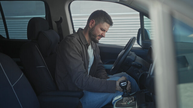 Man in car holds gearshift and leans forward inspecting the center console in building; contemplation.