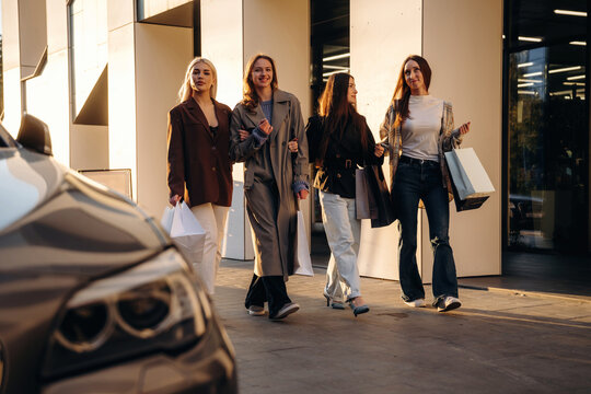 Four women walking together beside a parked car outside a modern building, carrying shopping bags in warm afternoon light, urban street scene