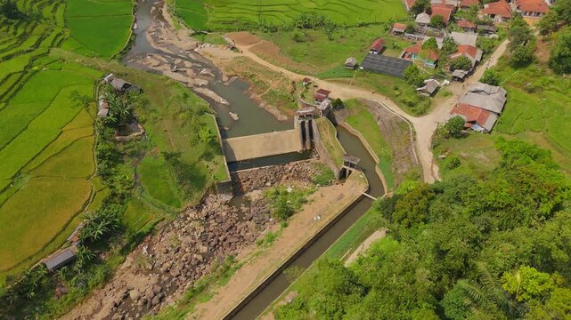 High altitude drone orbiting shot circling a concrete river dam, irrigation sluice gate, and surrounding countryside settlement