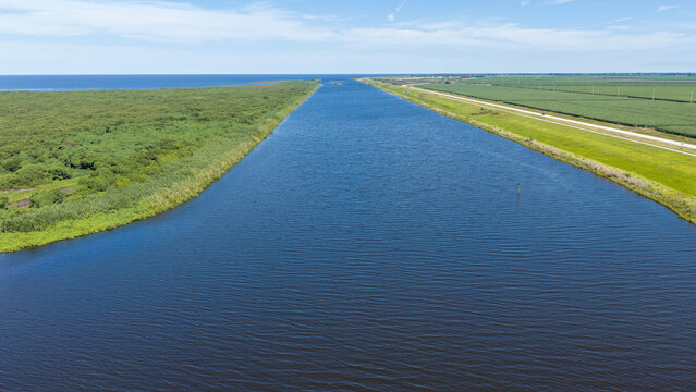 Aerial view of the canal leading into Lake Okeechobee, flanked by lush green vegetation and agricultural fields under a clear blue sky in Pahokee, Florida, United States.