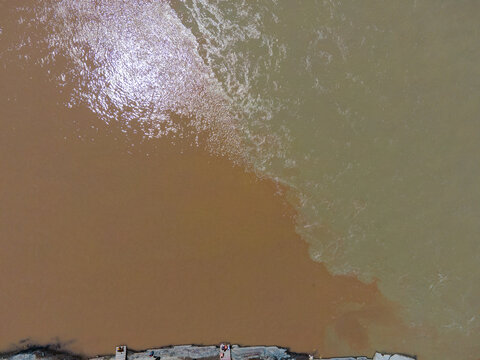 Aerial view of the confluence of the Zanskar and Indus rivers showing two distinct water colors meeting along a rocky riverbank with people visible in Ladakh Division, Ladakh, India.