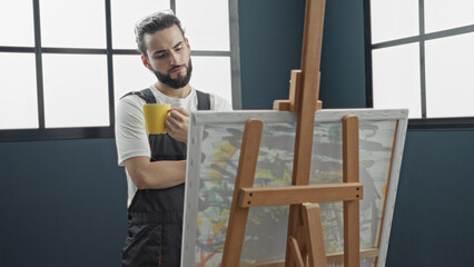 Man painter in studio wearing apron holds yellow mug and studies canvas on wooden easel with focused gaze  quiet contemplation. © Krakenimages.com