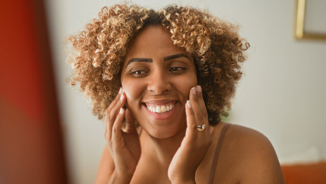 Woman with curly hair pressing cheeks and smiling into mirror in bedroom while wearing bra strap and ring on finger; self care joy.