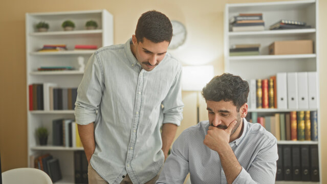 Two men at laptop, standing partner puts hand on seated colleague's shoulder while they review documents in an office building; support teamwork focus.