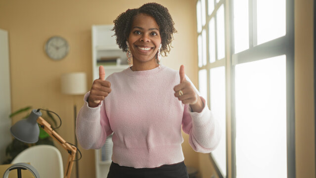 Young african american woman showing thumbs up with visible hands near desk lamp in studio; optimism.