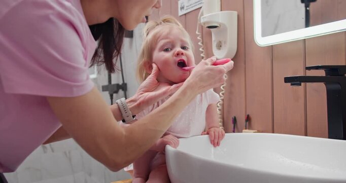 Mother brushing little daughter teeth by bathroom sink during morning routine. Daily oral hygiene from early childhood