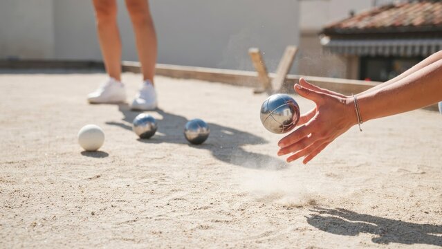 A hand releases a silver petanque ball on a sunny outdoor gravel court. Ideal for leisure activity and active lifestyle campaigns.