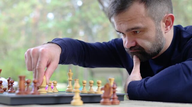 The man looks at the chessboard with concentration. He analyzes the situation on the board and makes a move with the knight. Playing chess in the city park. Slow motion 4K footage.