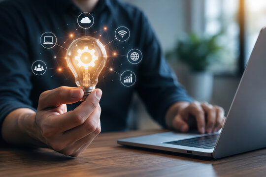 Hand holding illuminated lightbulb with gear icon above laptop in workspace representing digital innovation and online technology inspiration for startup ideas and business creativity