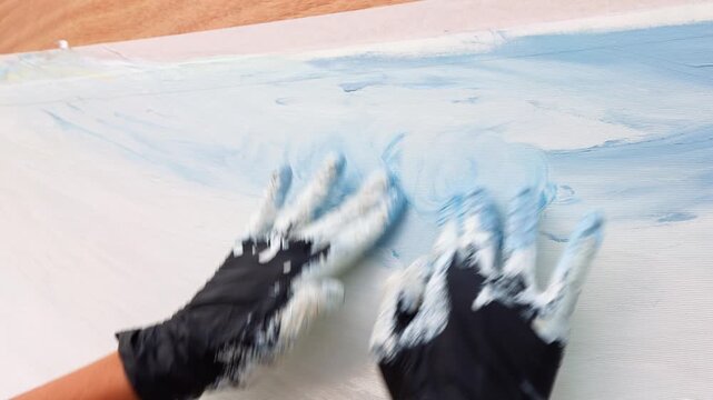 Close up view of artist hands wearing black protective rubber gloves covered in white and blue paint, applying smooth strokes directly onto a large white canvas to create modern abstract artwork