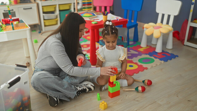 Woman stacking colorful blocks with little girl on foam playmat inside a building; playful learning.