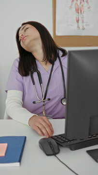 Woman in lavender medical scrubs with stethoscope at computer tilting head back and stretching neck at desk in building; fatigue stress recovery.