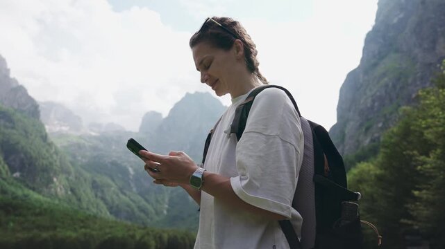 Young female traveler with a backpack using a smartphone while standing in a scenic mountain meadow, capturing mobile connectivity, navigation and modern adventure travel in nature.