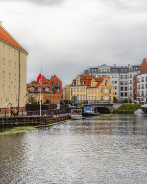 Gdansk, Pomeranian Voivodeship, Poland - 05 September 2026: View of the Motlawa River waterfront with historic buildings, a small bridge, and boats moored along the quay under a cloudy sky.