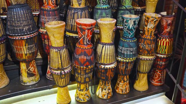 A vibrant collection of traditional djembe drums, each hand-painted with bold African tribal patterns in red, blue, yellow, and orange tones, displayed together on a market shelf.
