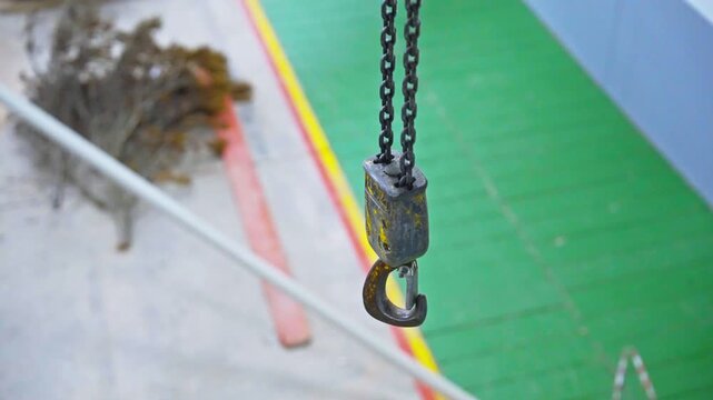 A close-up shot of a weathered metal chain hoist hook suspended from a double chain, with a softly blurred industrial background in green, blue, and orange tones.