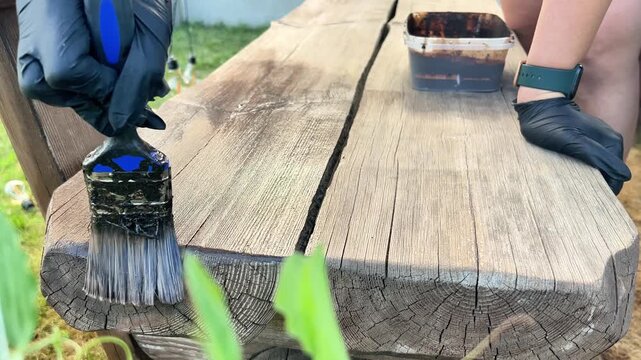Close-up of a person wearing black gloves applying protective dark stain to a wooden bench with a brush. Ideal for DIY, home improvement, and woodworking content.