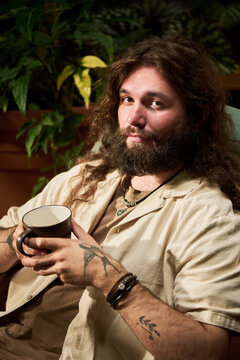 Vertical portrait of young adult Caucasian man with beard holding cup and relaxing indoors. Lifestyle visual for beverage branding and wellness marketing campaigns