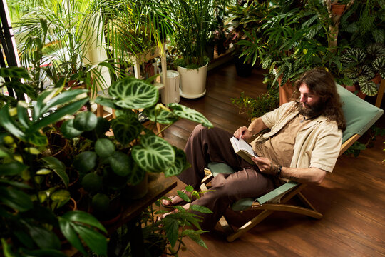 High angle shot of Caucasian young adult man reading book while sitting surrounded by houseplants. Concept of mental wellbeing, leisure time and eco lifestyle advertising