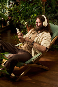 Vertical shot of young adult Caucasian man wearing headphones using smartphone while resting on deck chair surrounded by houseplants. Imagery suitable for audio streaming and wellbeing apps