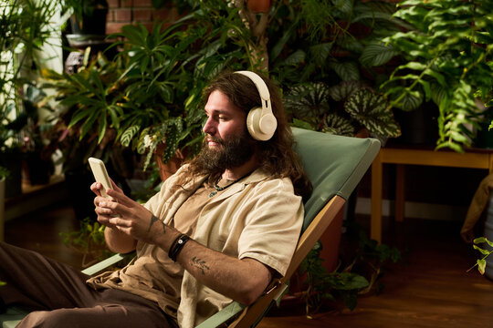 Young adult Caucasian man wearing headphones using smartphone while relaxing in chair surrounded by houseplants. Concept for audio streaming apps and digital wellness marketing