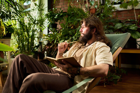 Young adult Caucasian man holding notebook and pencil while resting among house plants, side view. Visual metaphor representing creative inspiration, mindfulness, and journaling advertising