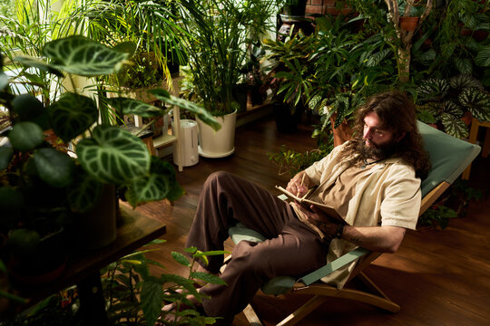 Young adult Caucasian man writing in notebook while sitting surrounded by houseplants. Visual represents mindfulness and creativity, suitable for mental health campaigns