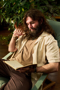 Vertical shot of young adult Caucasian man sitting, writing notes in notebook, holding pencil thoughtfully. Image suited for creative writing courses and mindfulness campaigns